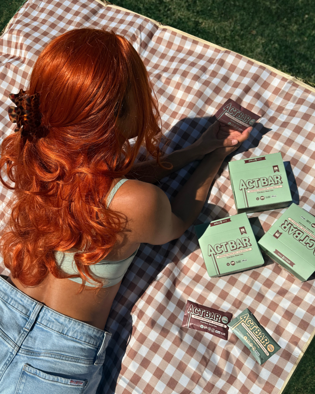Person with red hair lying on a checkered blanket with ACT BAR products around them.